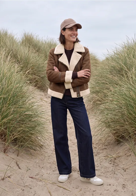 Frau in brauner Lammfelljacke, dunkler Hose und Mütze steht am Strand vor Dünengras.