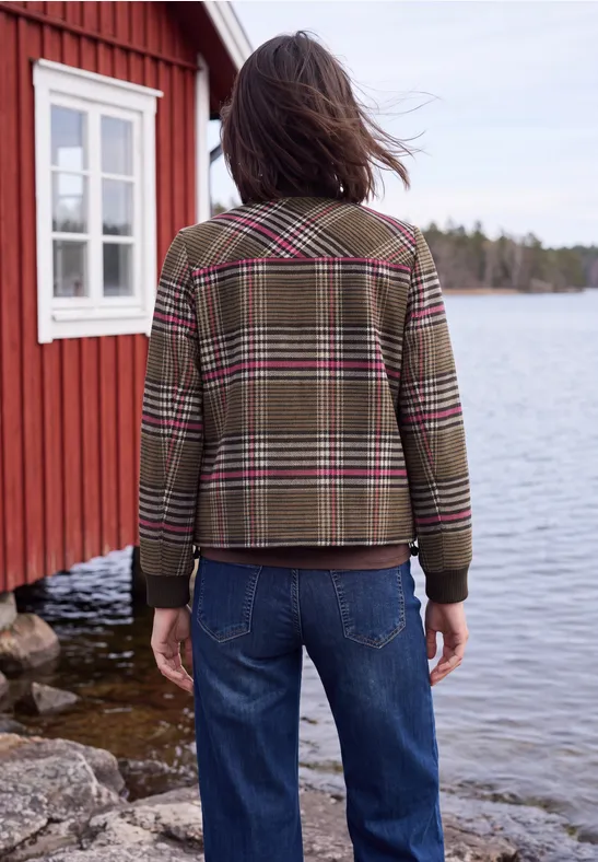 Rückansicht einer Person in karierter Jacke und Jeans, die am Ufer vor einem roten Holzhaus an einem See steht.