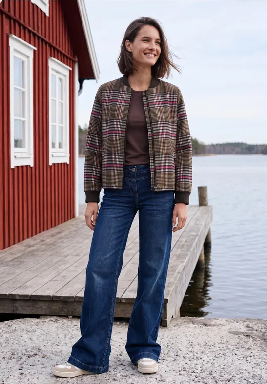 Frau in Karojacke und Jeans steht auf einem Steg neben einem roten Haus am Wasser und lächelt.