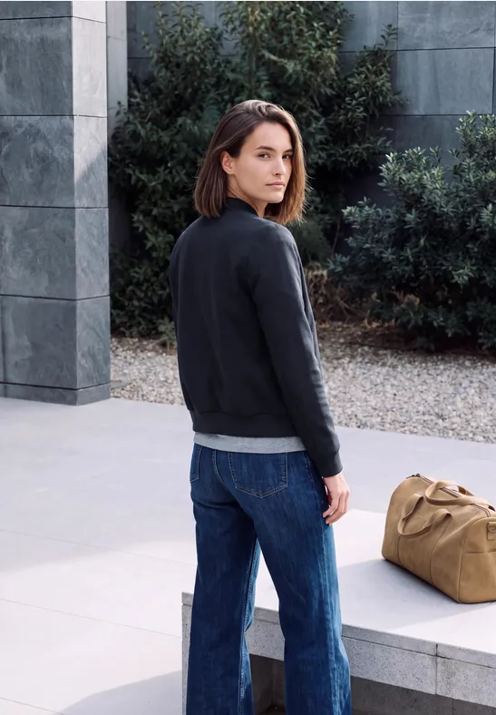 Een vrouw in jeans en trui staat bij een betonnen gebouw, kijkt achterom, naast haar ligt een sporttas.