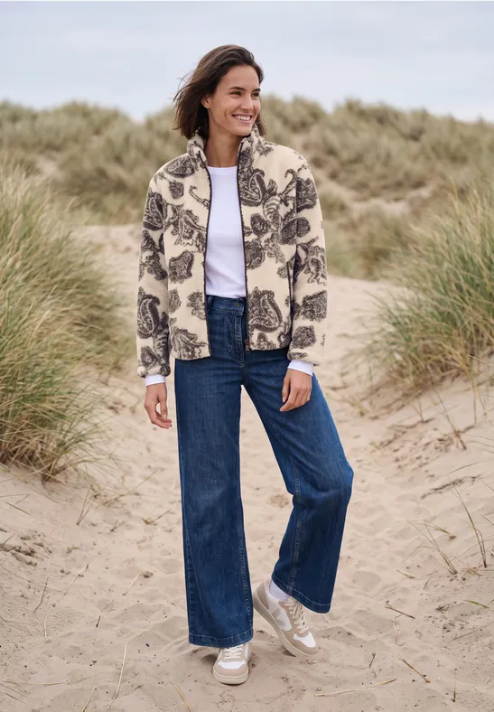 Frau in gemustertem Cardigan, weitem Jeans und Sneakern am Strand