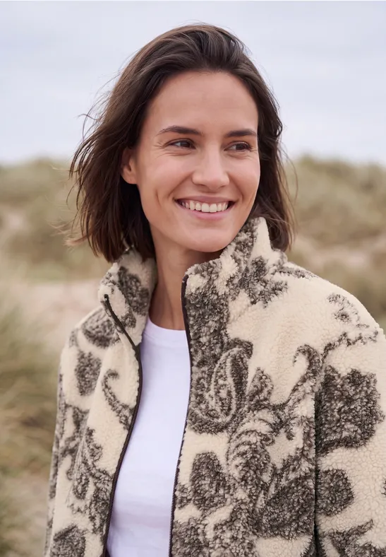 Frau lächelt am Strand, trägt gemusterte Fleecejacke, Dünen im Hintergrund.