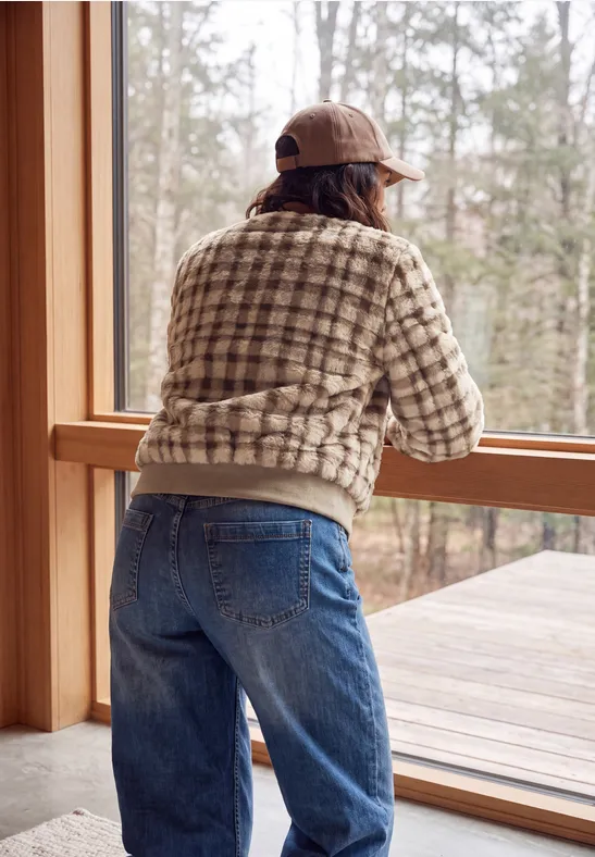 Person im karierten Pullover, Jeans und Cap lehnt am Holzfenster und schaut hinaus in den Wald.