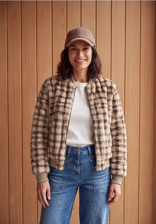 Eine Frau in einer beige-karierten Jacke, weißem T-Shirt, blauen Jeans und Beanie lächelt vor Holzpaneelen.