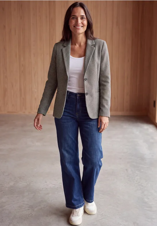 Frau in grauem Blazer, weißem Top, blauen Jeans und weißen Sneakers steht in modernem Raum mit Holzverkleidung und lächelt.