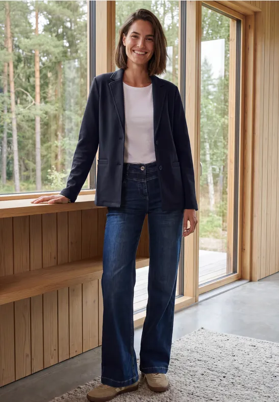 Frau mit dunkelblauer Jacke und Jeans lehnt lächelnd an einer Holzfensterbank mit Waldblick.