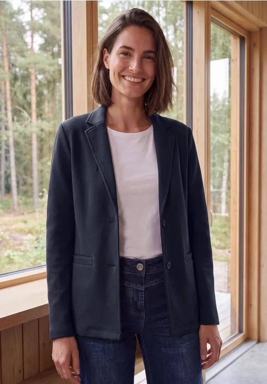 Eine lächelnde Frau in Blazer und Jeans steht vor einem Fenster mit Blick auf den Wald.