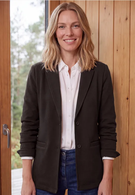 Eine lächelnde Frau mit blondem Haar, schwarzem Blazer, weißem Hemd und Jeans vor einer Holzverkleidung.