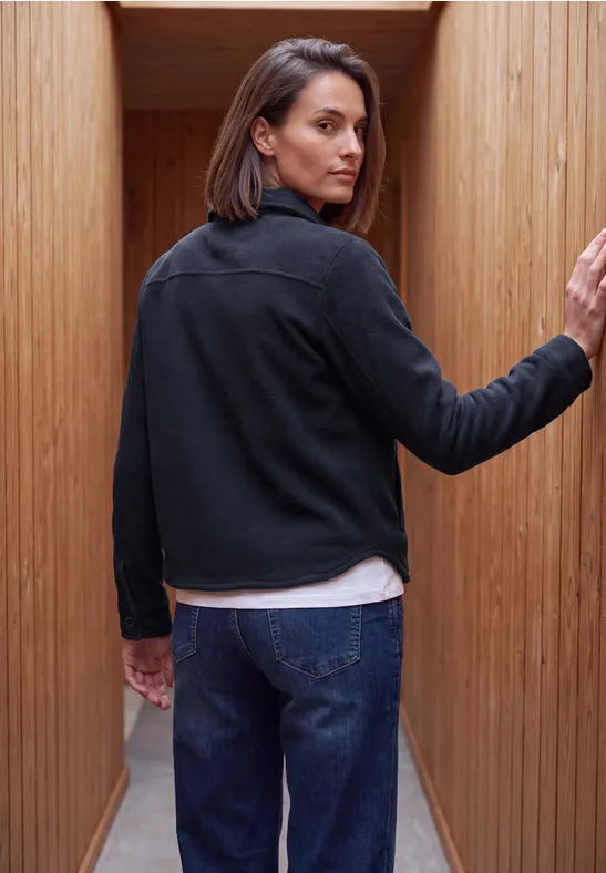 Eine Frau in schwarzer Jacke und Jeans blickt über die Schulter in einem schmalen Holzgang.