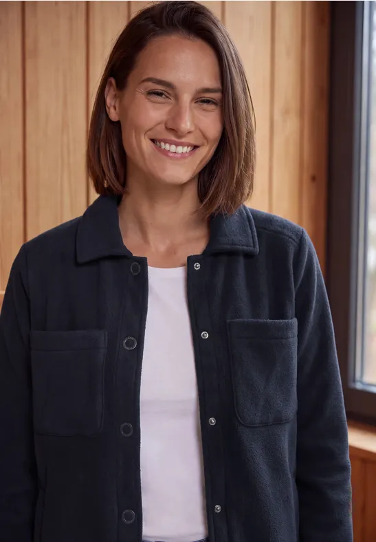 Eine lächelnde Frau in weißem T-Shirt und dunkelblauer Hemdjacke steht vor einer Holzwand.