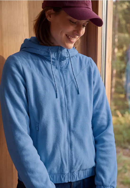 Jonge vrouw in een blauw capuchonvest en pet die glimlacht bij een raam met boszicht.