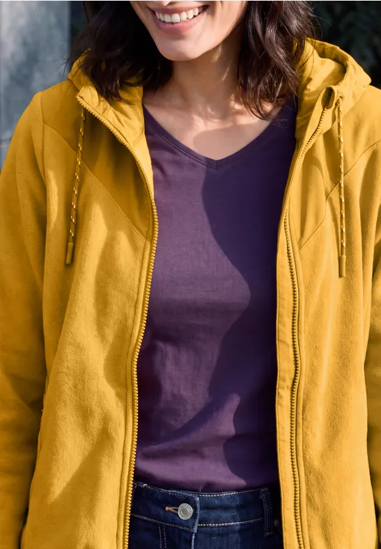 Frau trägt gelbe Kapuzenjacke über lila T-Shirt, nur Oberkörper und leichtes Lächeln sichtbar.