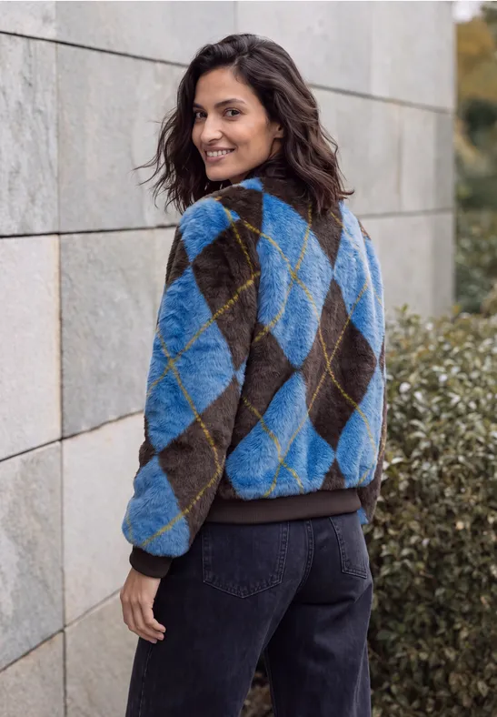 Eine Frau in einem karierten Pullover blickt über die Schulter vor Mauer und Sträuchern.