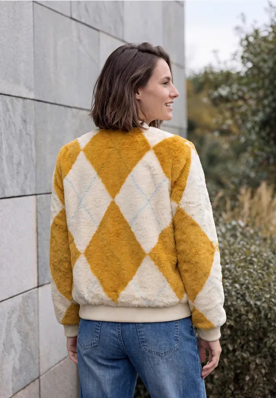 Eine Frau steht seitlich an einer Steinwand, trägt eine gelb-weiße Rautenmuster-Jacke und Jeans.