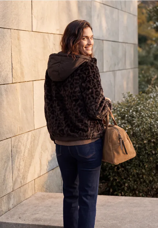 Une femme avec un sac marron et un sweat à capuche sombre, devant un mur, regardant par-dessus son épaule.
