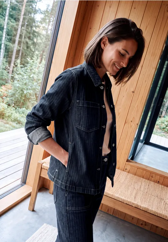 Frau im dunklen Denim-Outfit steht lächelnd am Fenster in einem hellen Holzraum.