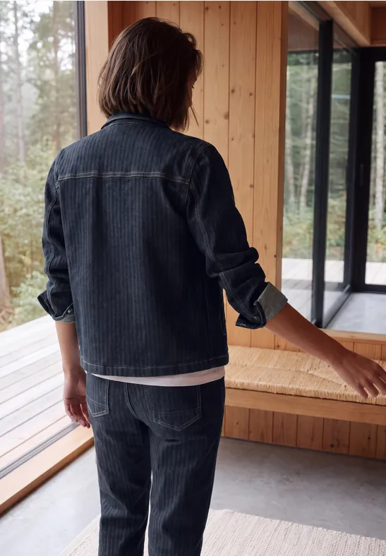 Person von hinten in Jeansjacke und Jeanshose steht in modernem Holzhaus mit großen Fensterfronten.