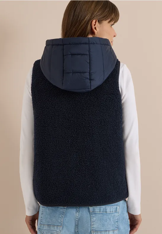 Rückansicht einer Person in weißem Langarmshirt und dunkelblauer, ärmelloser Fleece-Weste mit Kapuze, blaue Jeans.