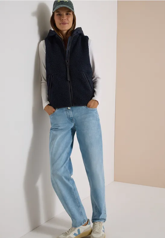 Frau in marineblauer Fleece-Weste, weißem Langarmshirt, hellblauen Jeans, weißen Sneakern und Cap lehnt in Ecke.