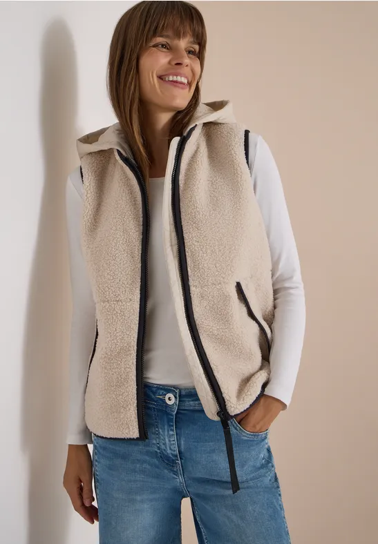 Vrouw in crèmekleurig fleecevest, wit shirt met lange mouwen en spijkerbroek, glimlacht ontspannen.
