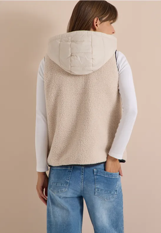 Achteraanzicht van vrouw in crèmewitte, gewatteerde bodywarmer met capuchon, wit shirt en blauwe jeans.