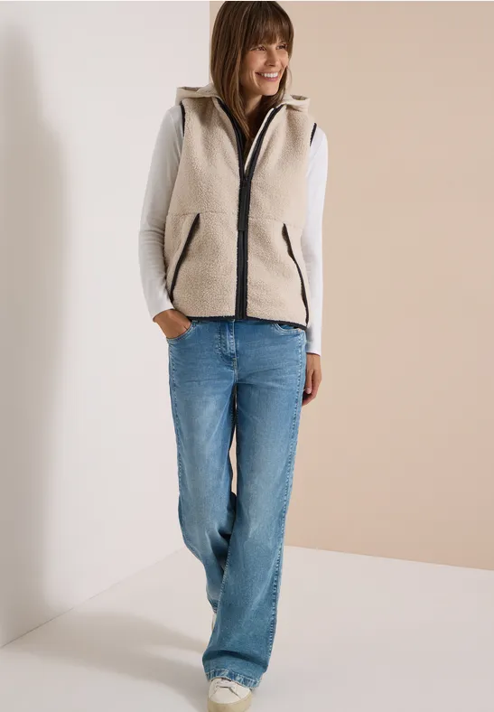 Vrouw in grijze fleecebodywarmer met capuchon, grijs shirt met lange mouwen en blauwe jeans, glimlachend, handen in zakken.