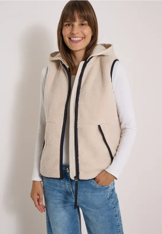 Vrouw lacht in lichtbeige fleecevest met ritssluiting, witte top en blauwe jeans.