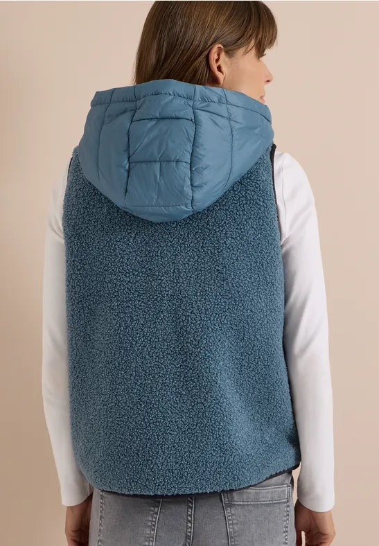 Frau von hinten in jeansblauer Teddyweste mit gesteppter Kapuze und weißem Langarmshirt.