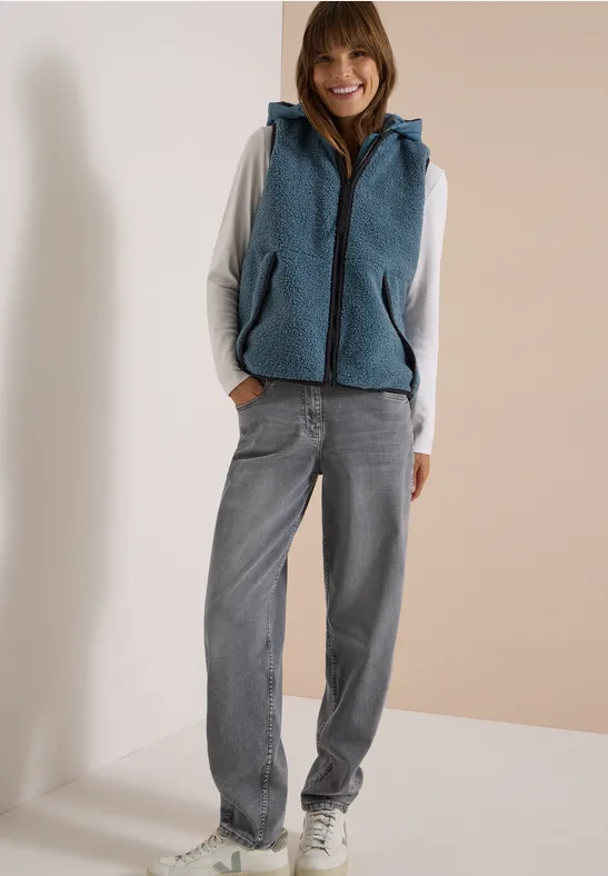 Eine Frau trägt einen grauen Hoodie, eine blaue Fleece-Weste und eine weite Jeans.