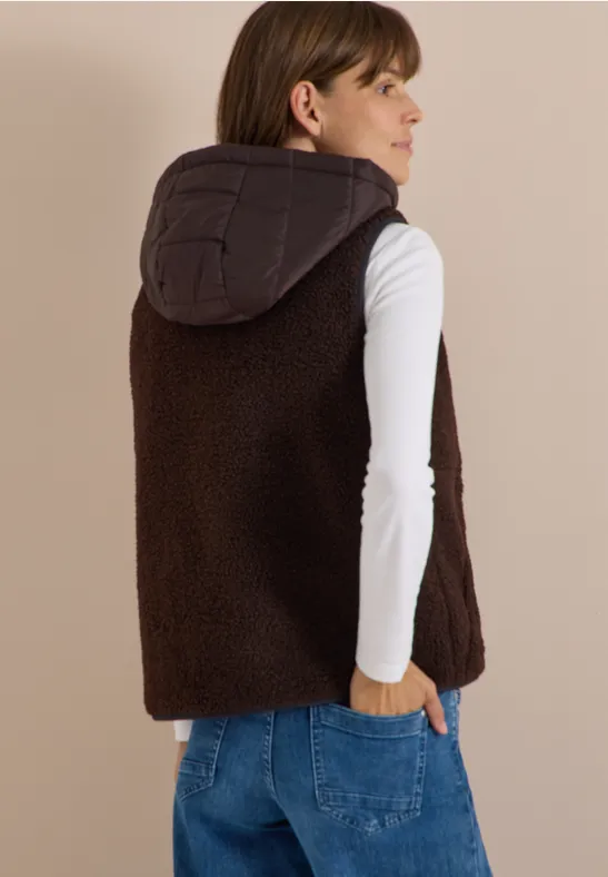 Vrouw van achteren met witte longsleeve, blauwe jeans en bruine teddyvest met capuchon.