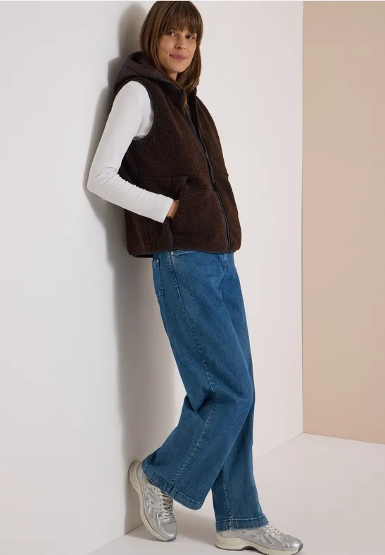 Vrouw leunt tegen muur, draagt bruine fleecevest met capuchon, witte trui, wijde blauwe jeans en grijze sneakers.