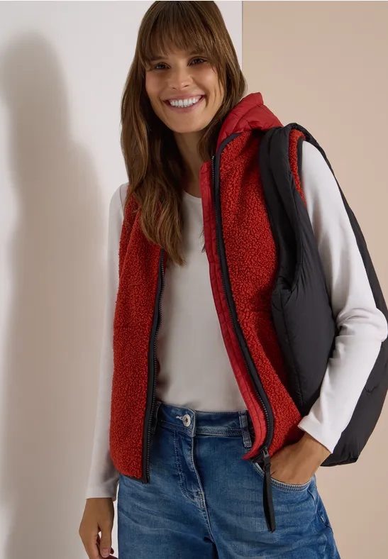 Lächelnde Frau in roter Steppweste mit Kapuze, weißem Langarmshirt und blauen Jeans.