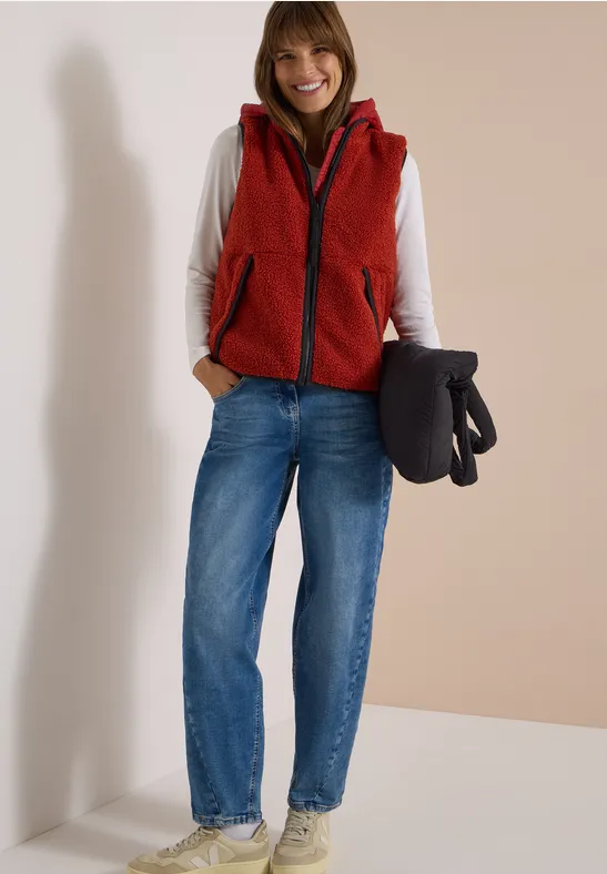 Eine lächelnde Frau trägt rote Weste, weiße Bluse, blaue Jeans und weiße Sneaker, hält schwarzen Mantel.