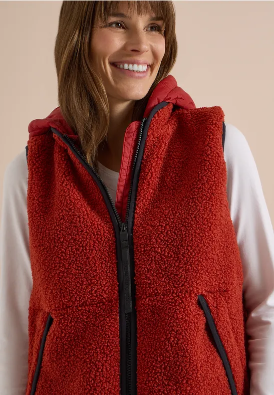 Lächelnde Frau in roter Fleece-Weste mit Kapuze und Reißverschlusstaschen über weißem Langarmshirt.