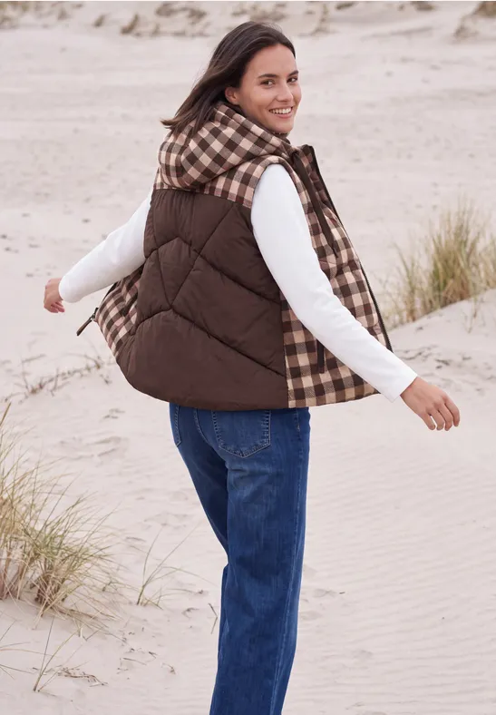 Eine lächelnde Person in brauner Weste und Jeans, die am Strand über Sanddünen spaziert.