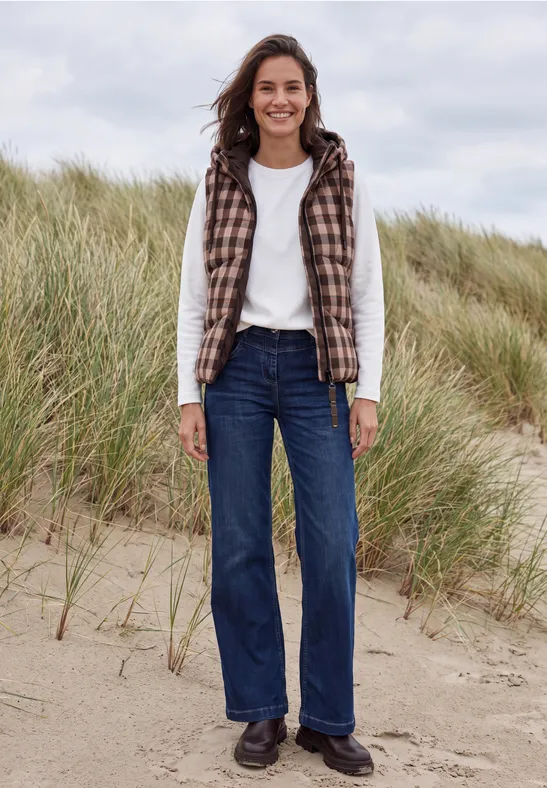Eine lächelnde Frau trägt Jeans, weißen Pullover und kariertes Gilet am sandigen Dünenstrand.