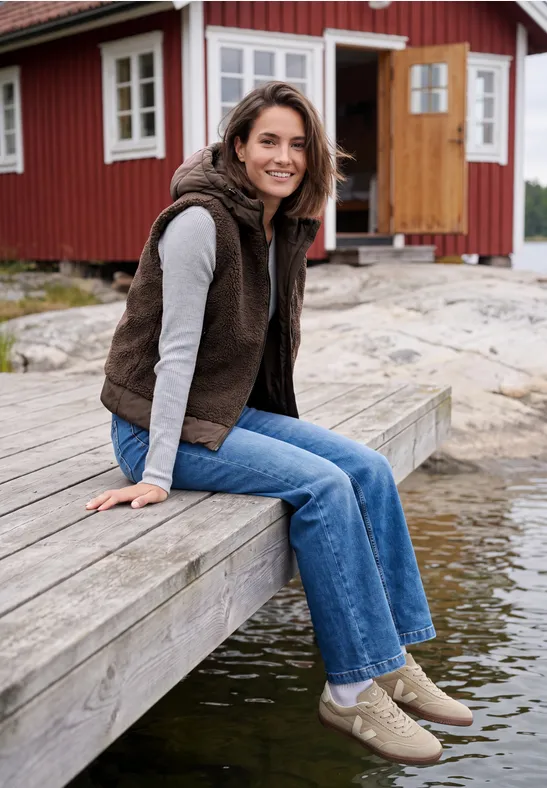 Eine Frau sitzt lächelnd auf einem Steg vor einem roten Holzhaus und blickt aufs Wasser.