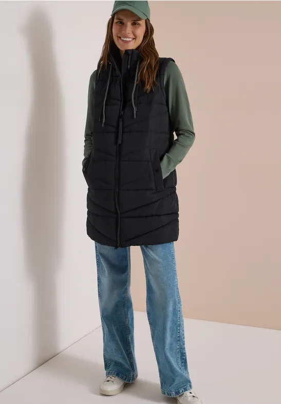Femme portant une longue veste matelassée noire, un sweat à capuche vert, un jean large et une casquette.