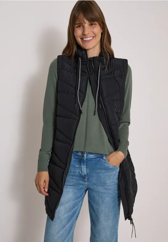 Femme portant une longue doudoune sans manches noire, pull vert et jean bleu, souriante sur fond blanc.