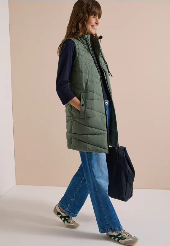 Femme en longue veste matelassée à capuche, top sombre, jean large, baskets et grand sac fourre-tout.