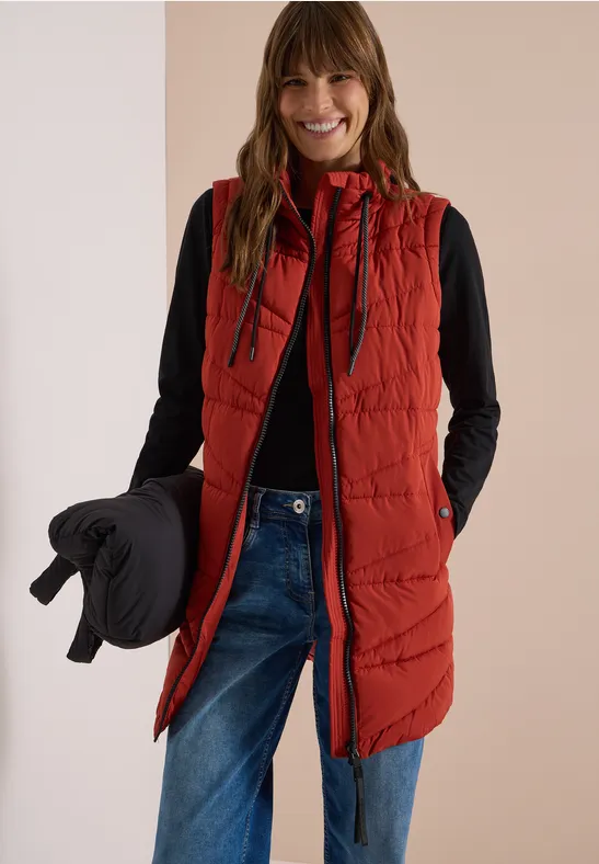 Femme souriante portant une veste sans manches rouge, un top noir et un jean, tenant une doudoune noire.