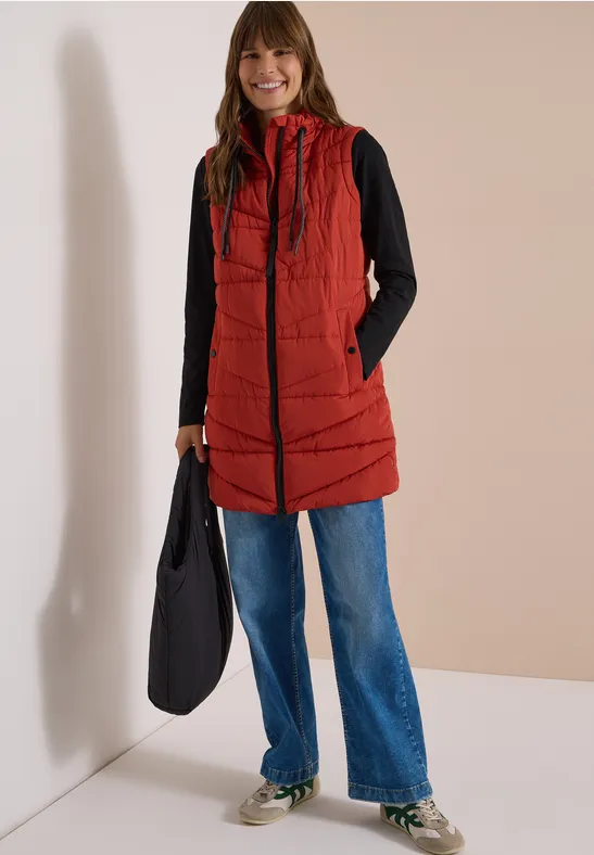 Femme souriante en doudoune longue rouge à capuche, pull noir, jean bleu et sac noir.