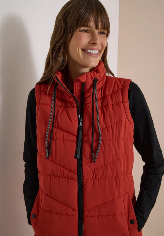 Femme souriante portant un gilet matelassé rouge à capuche sur un top noir à manches longues.