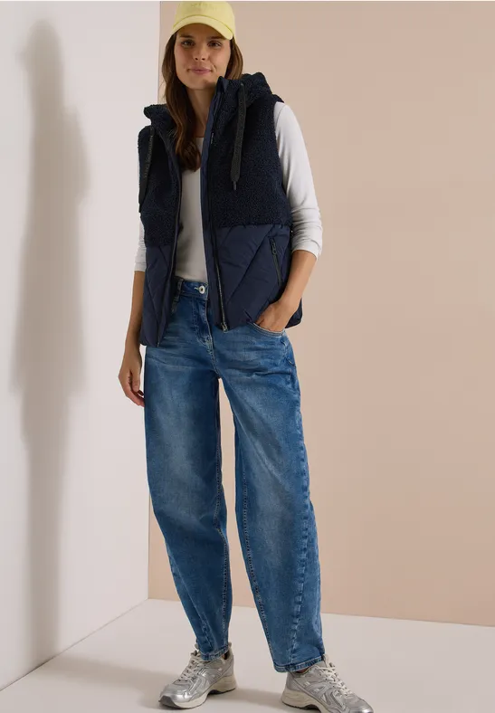 Femme en tenue décontractée : jean ample, t-shirt à manches longues blanc, doudoune sans manches, casquette et baskets.