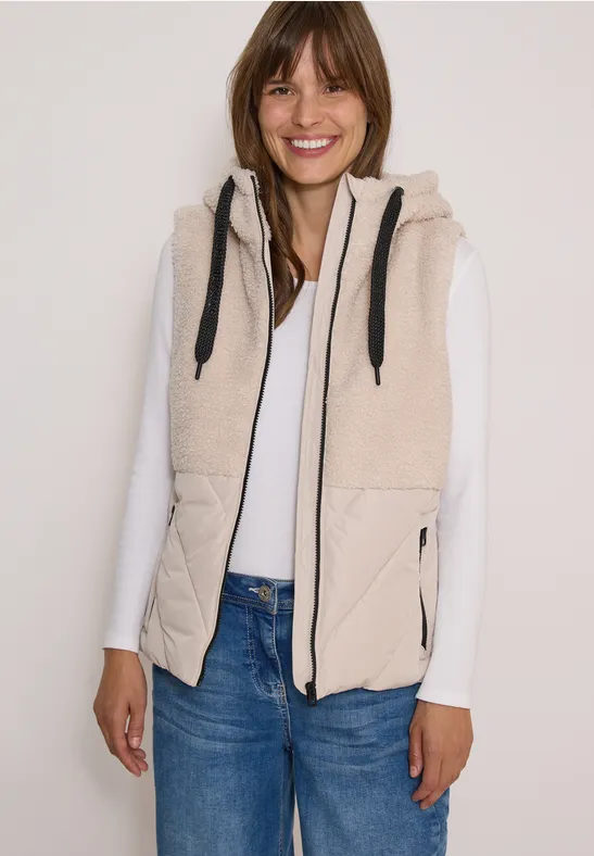 Femme porte un gilet beige à capuche sur un t-shirt blanc et un jean