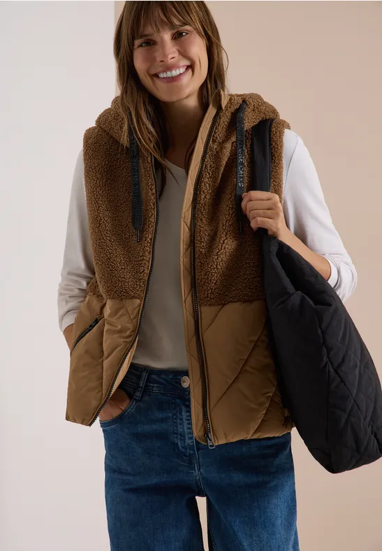 Lächelnde Frau in brauner Fleece-Steppweste, beigem Pullover, Jeans und schwarzer Tasche vor hellem Hintergrund.