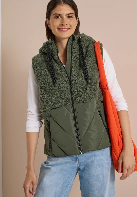 Une femme porte une doudoune sans manches olive à capuche, un tee-shirt blanc à manches longues et un tote bag orange.