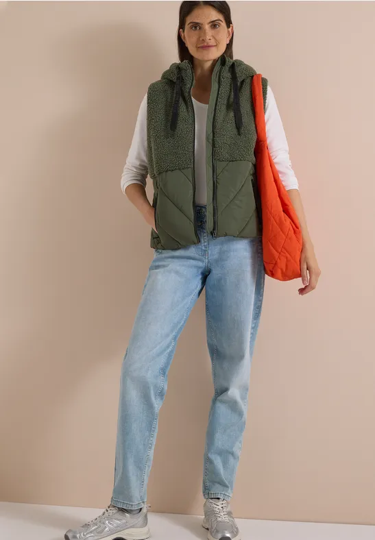 Femme en gilet matelassé kaki sur t-shirt blanc, jean et baskets, tenant un sac orange devant un mur beige.