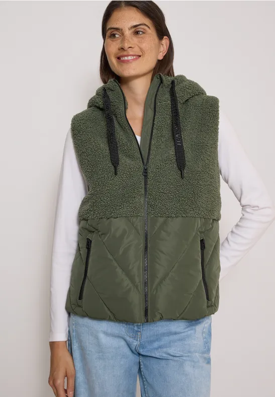 Femme en t-shirt manches longues blanc et jeans porte une veste sans manches en polaire verte à capuche zippée.