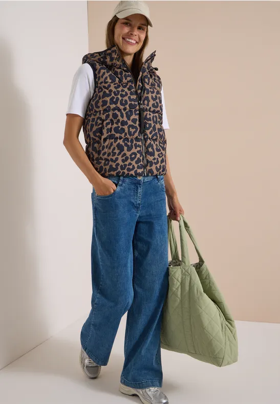 Frau in leo-gemusterter Weste, weißem Shirt, weitem Jeans, Sneakern und großer grüner Tasche.